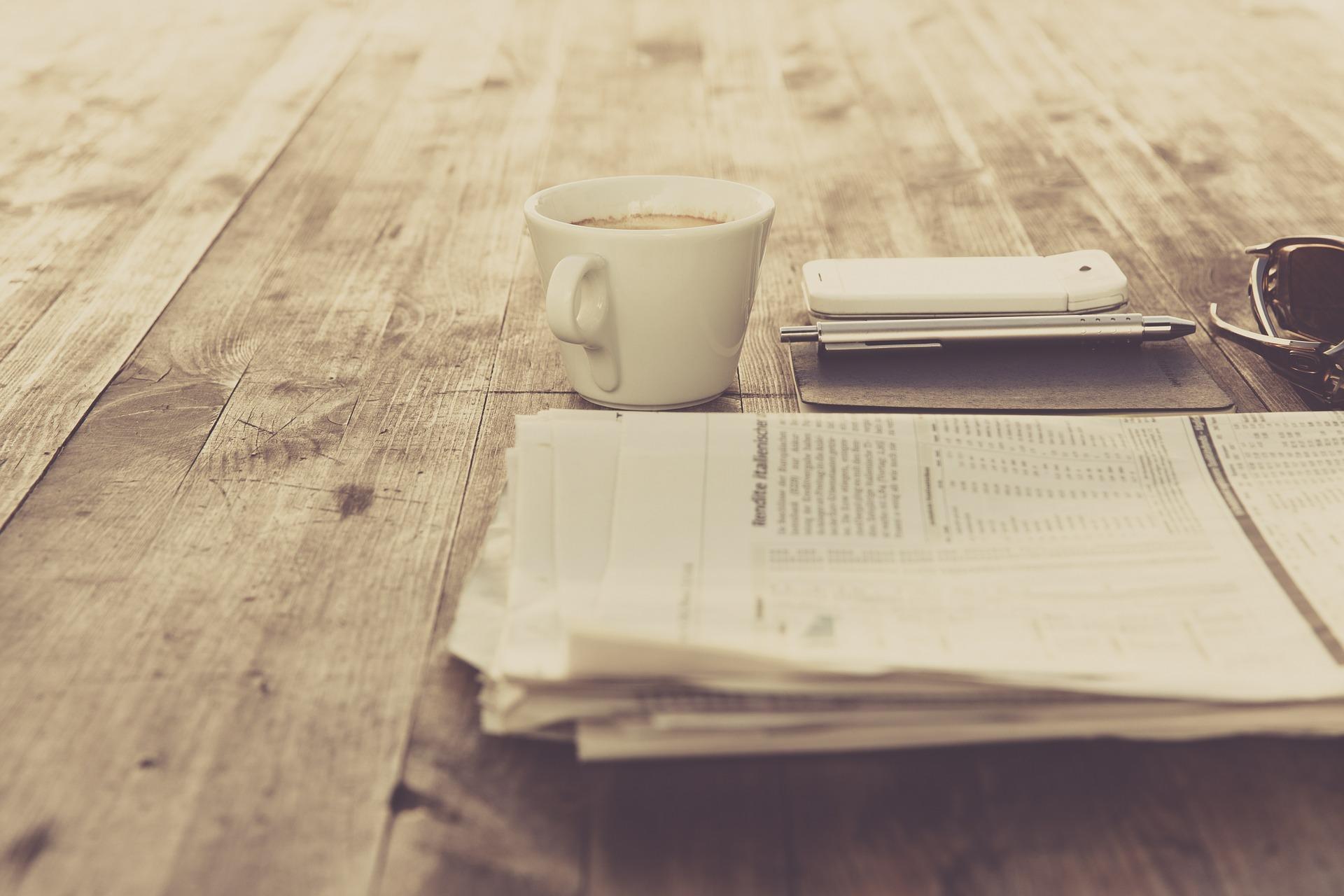 Zeitung neben Kaffeetasse und Handy auf Holztisch.