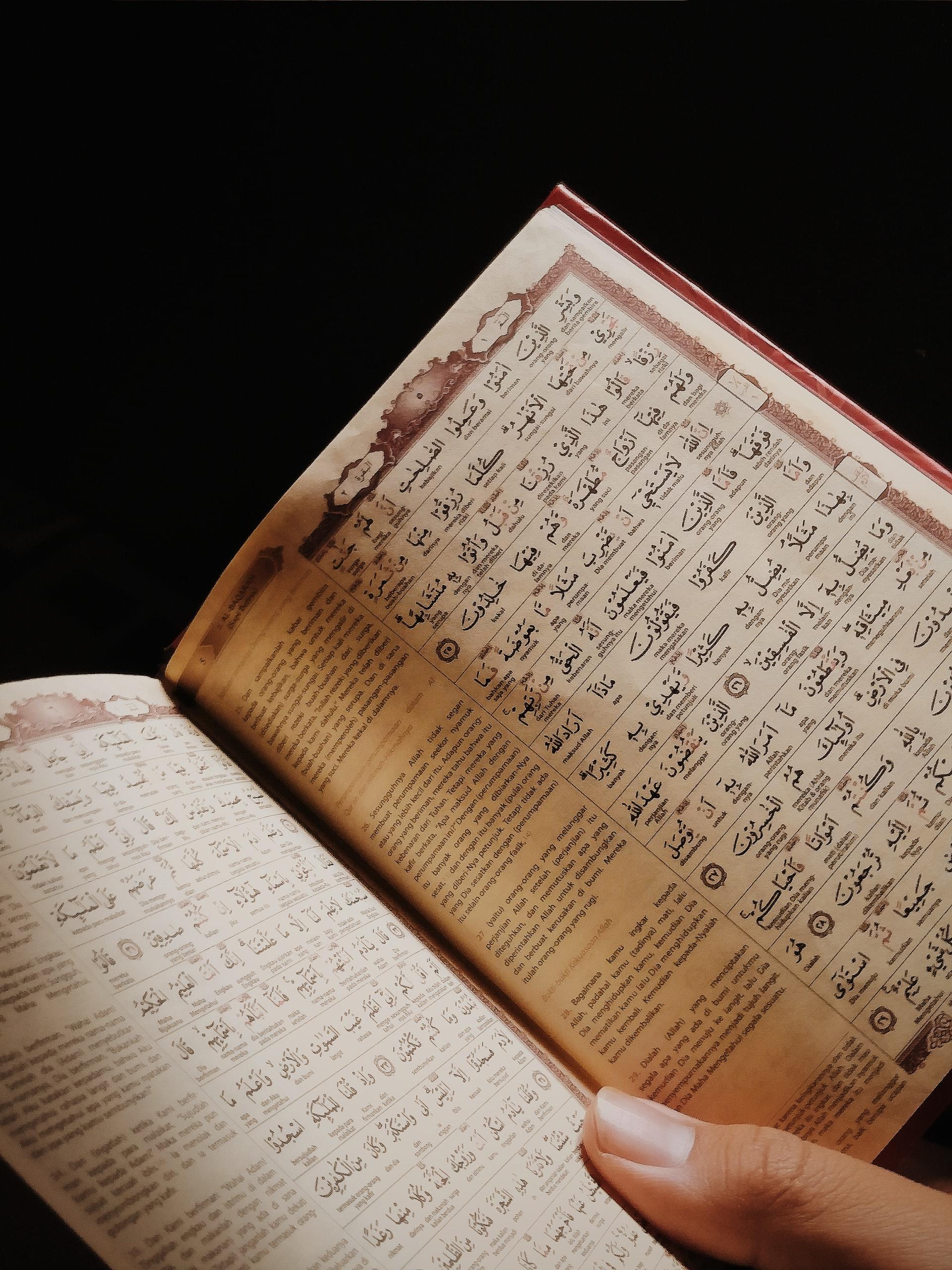 Ein aufgeschlagenes Buch weist pro Seite jeweils eine linke Spalte mit Texten in in römische Buchstaben übertragener arabischer Sprache und eine rechte Spalte mit pro Zeile mehreren einzelnen Wörtern in arabischer Schrift und darunter jeweils ebenfalls in römischen Buchstaben verfassten arabischsprachigen Übertragungen dieser Wörter auf.