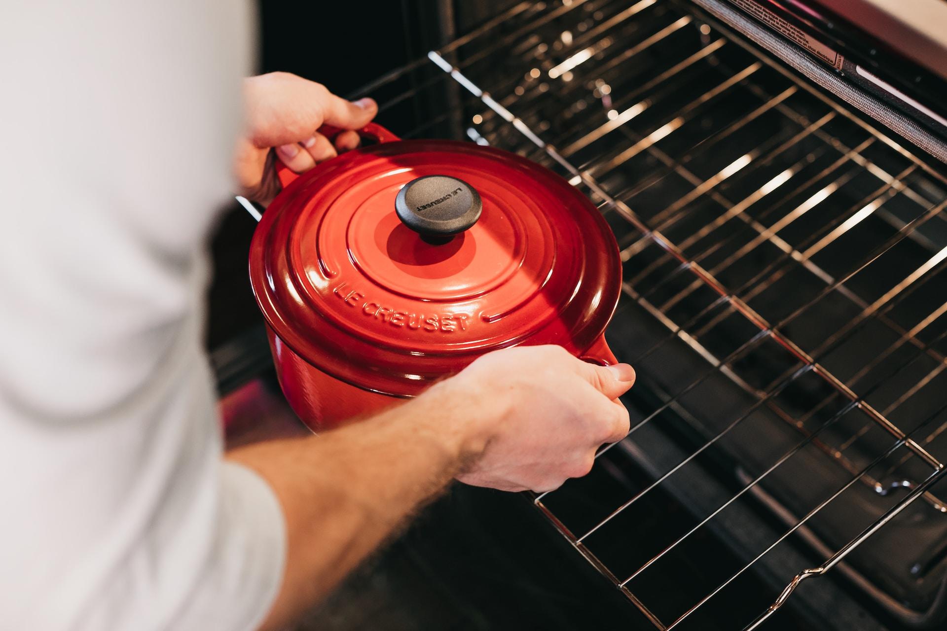 Roter Gusseisentopf mit Coc au Vin wird wird von Mann in weißem Pullover in den Backofen geschoben.