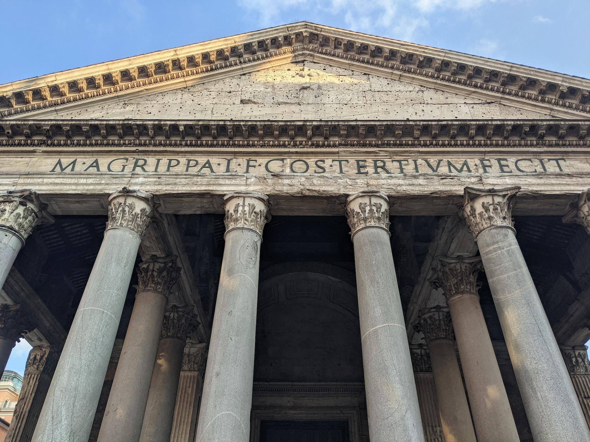 Portal von römischem Gebäude mit lateinischen Wörtern über den Säulen.