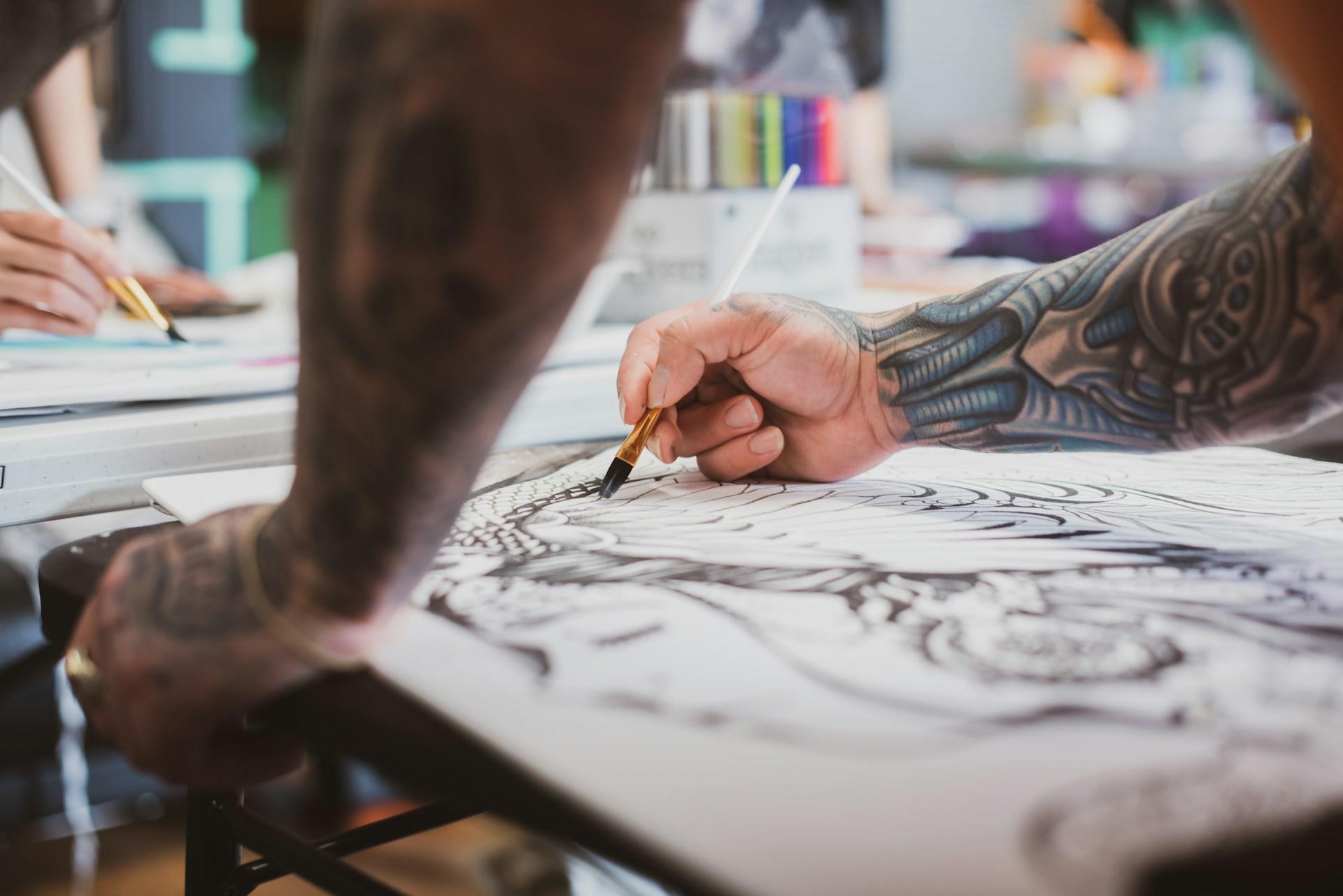 Ein tättowierter Mann steht über einem Zeichenblock und malt mit einem Pinsel eine Tattoovorlage