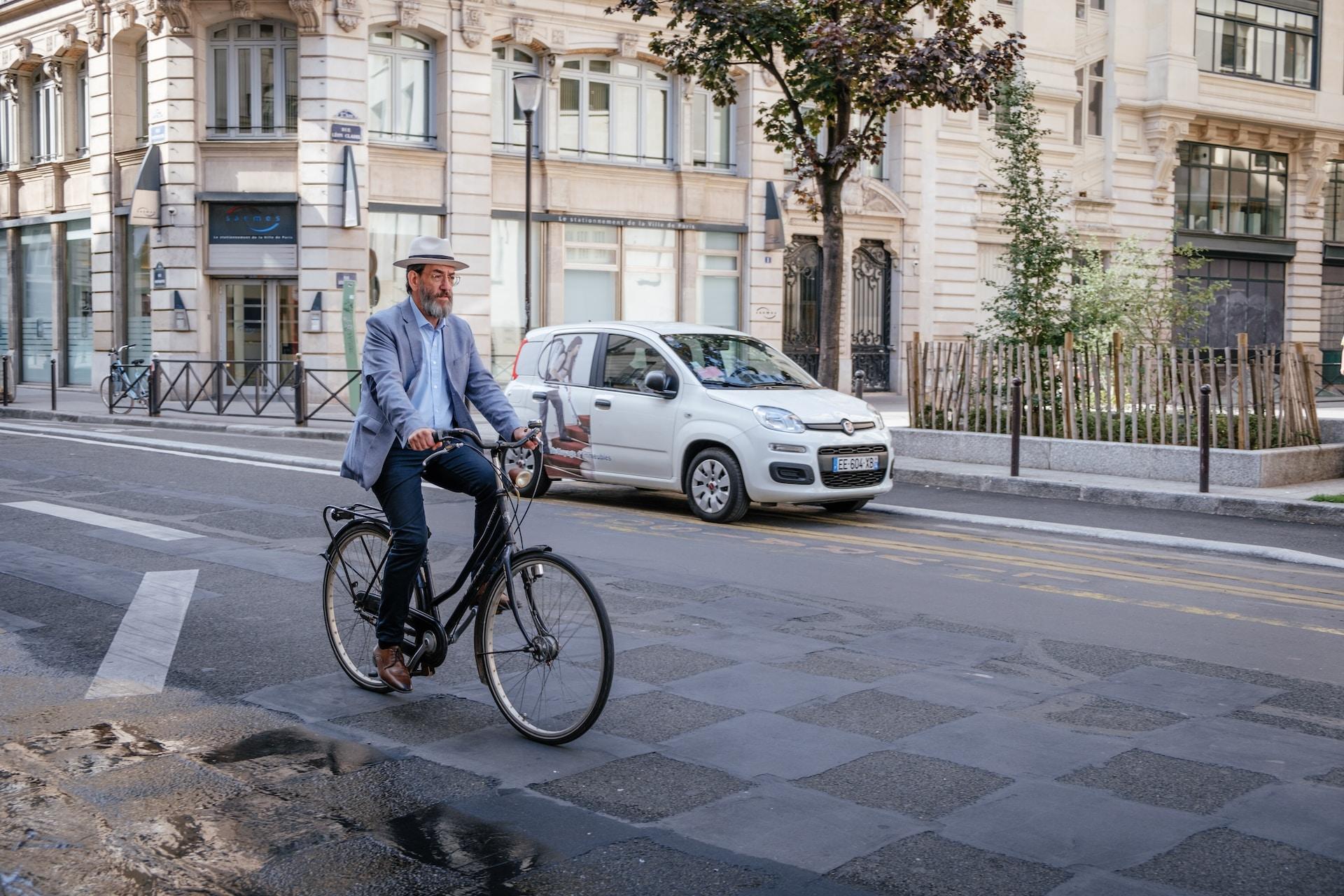 Älterer Mann fährt mit Rad auf einer Straße von links und im Hintergrund steht ein weißes Auto.