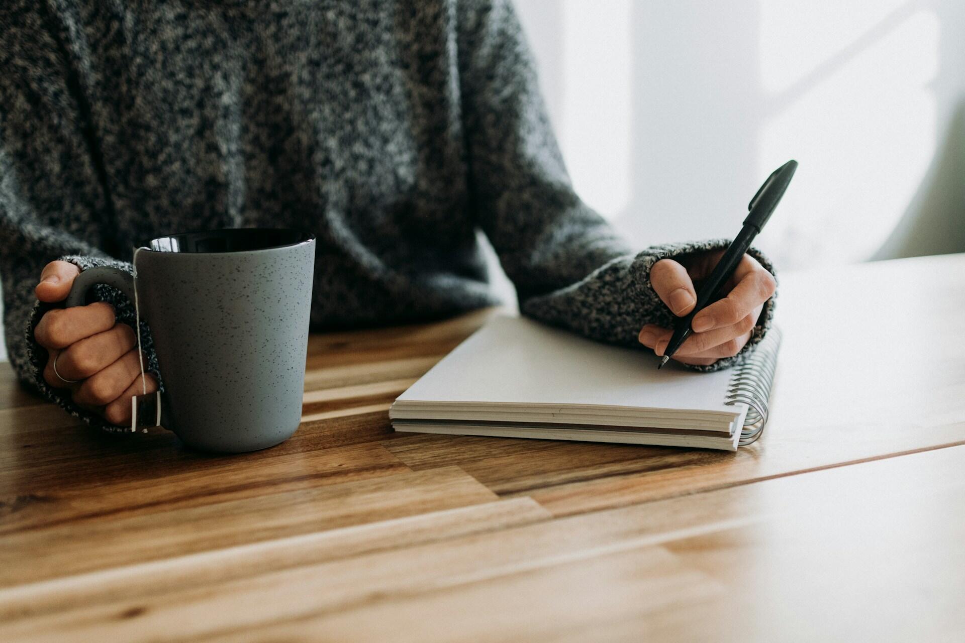 Frau schreibt mit Stift in Block mit Kaffeetasse in der Hand.