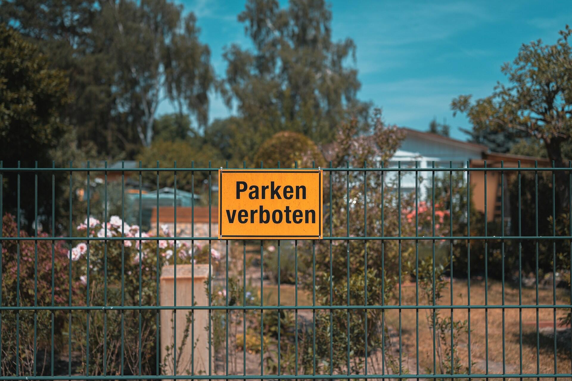 Gelbes Schild mit schwarzer Schrift an grünem Zaun vor Bäumen.