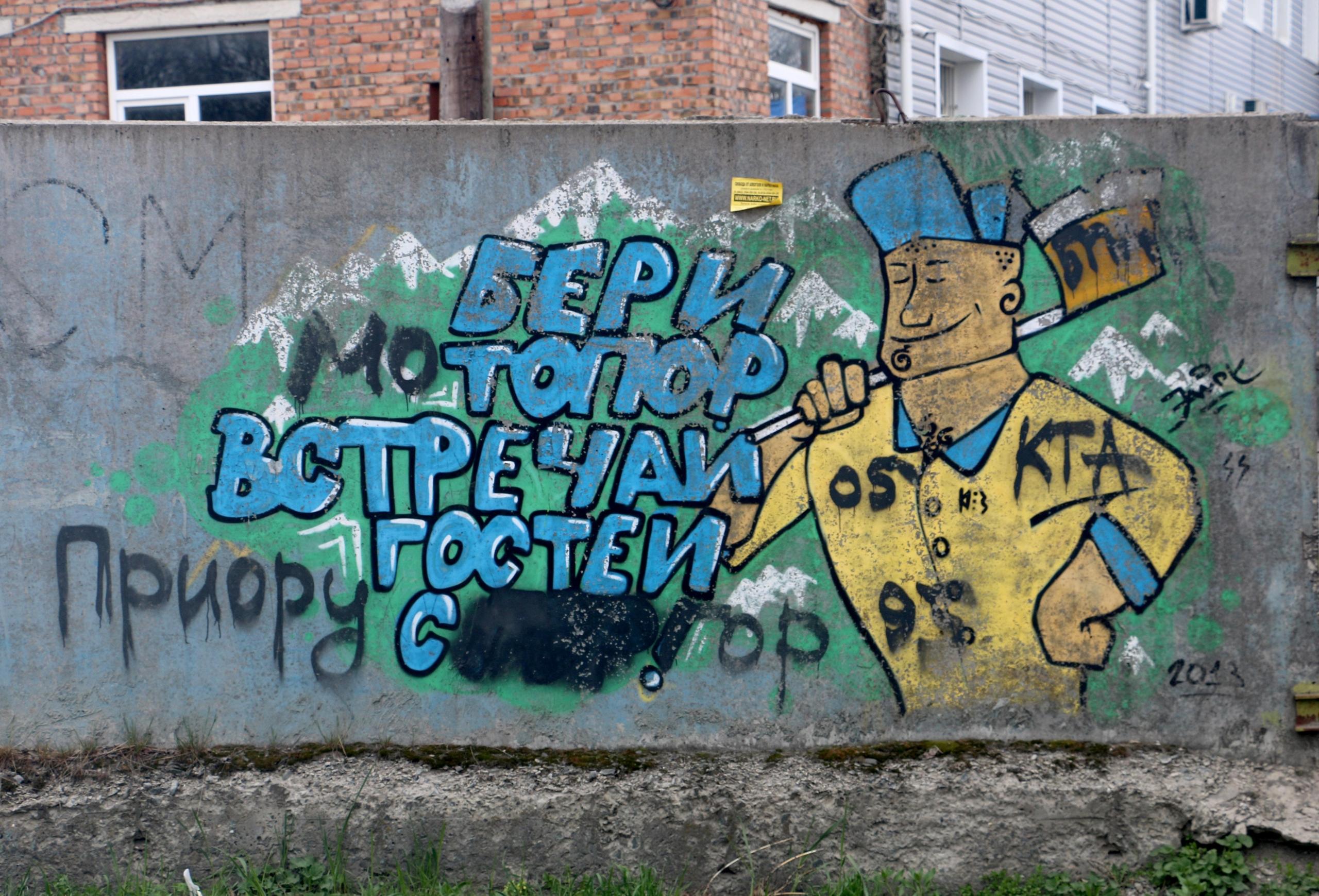 Ein Graffiti an einer Wand mit russischer Schrift und einem daneben gesprayten Bauarbeiter, der eine Axt hält und lächelt.