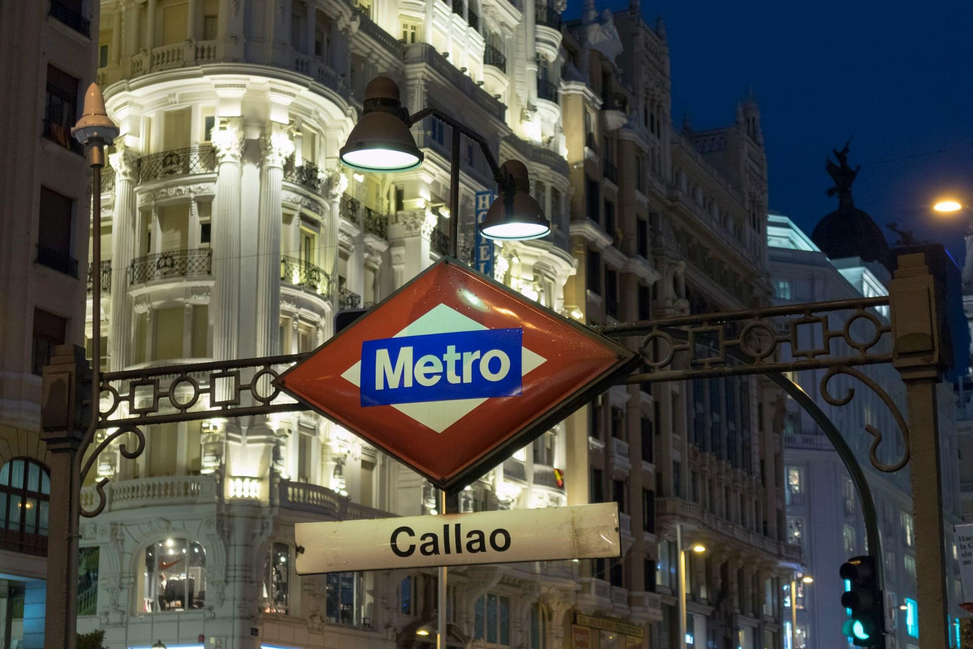 Ein Metro Schild bei Nacht mit hell erleuchteten alten Gebäuden im Hintergrund in der Spanischen Hauptstadt Madrid.