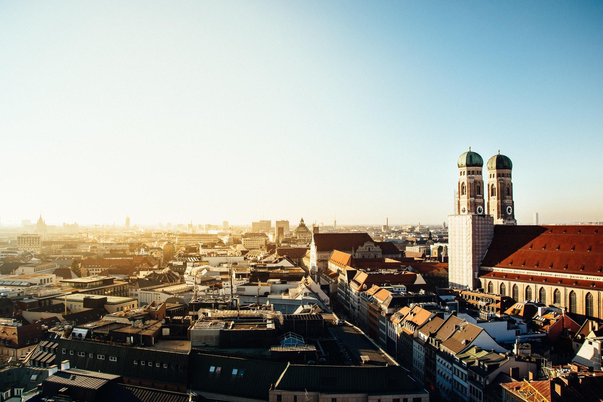 Ein Panoramafoto über die Dächer von München bei blauem Himmel, rechts sind die zwei Türme der Frauenkirche zu sehen.
