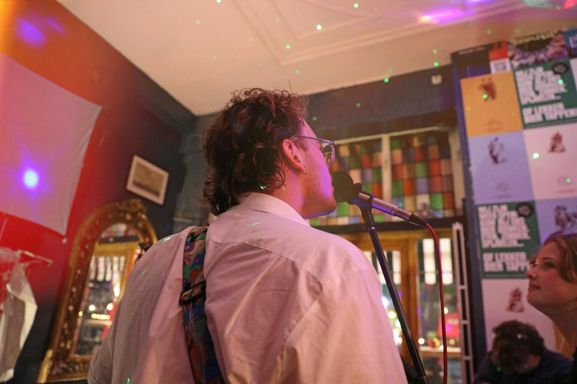 Mann mit Hemd steht in Bar und singt in Mikrofon.