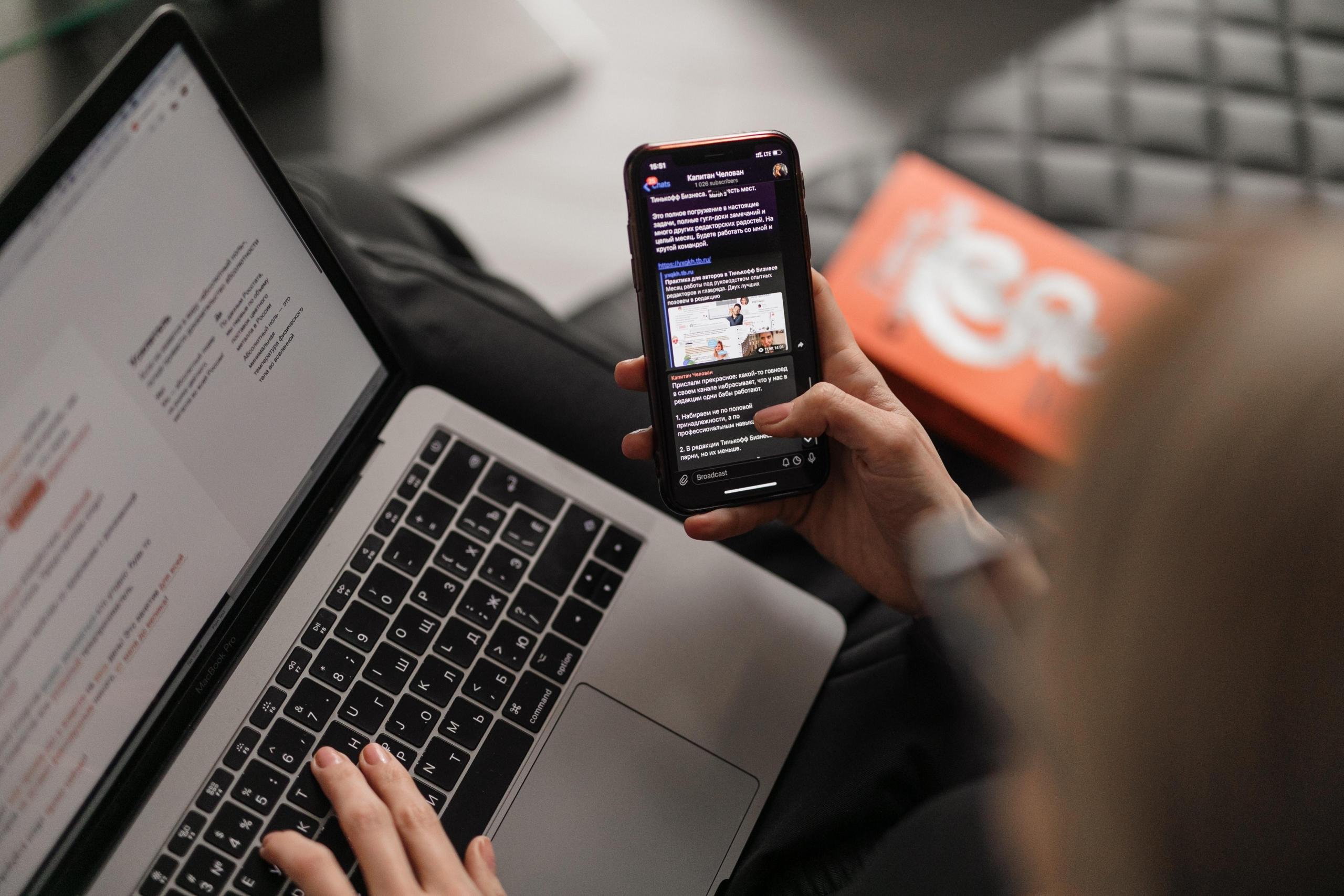 Eine Person hat einen Laptop auf dem Schoß und ein Handy in der Hand