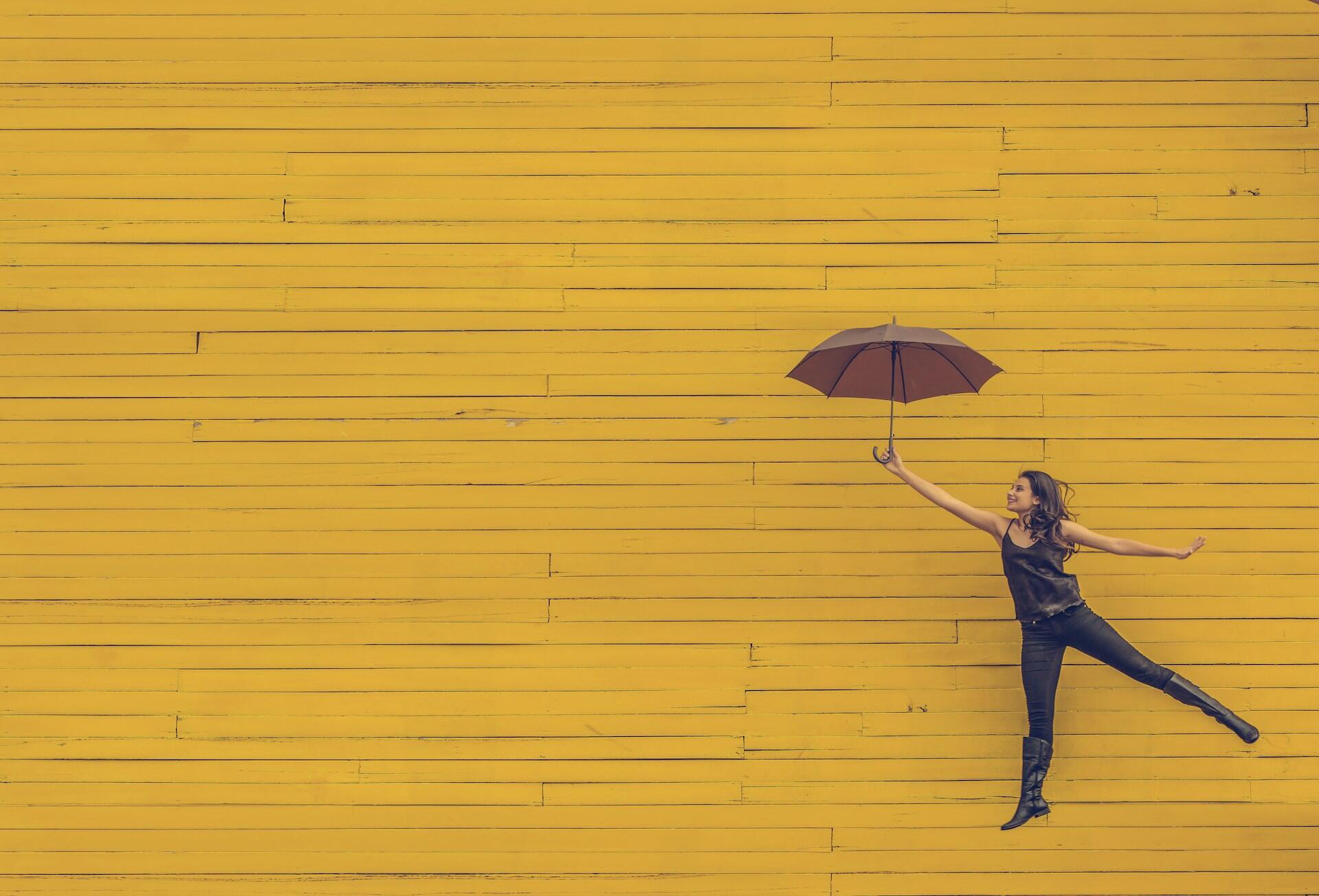 Frau mit Regenschirm springt vor gelber Wand.