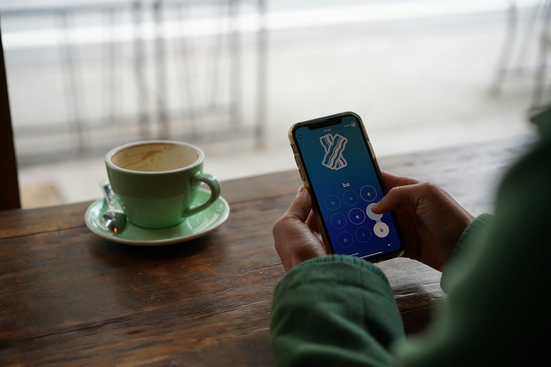 Zwei Hände tippen auf einem Smartphone und daneben steht eine Tasse auf dem Tisch