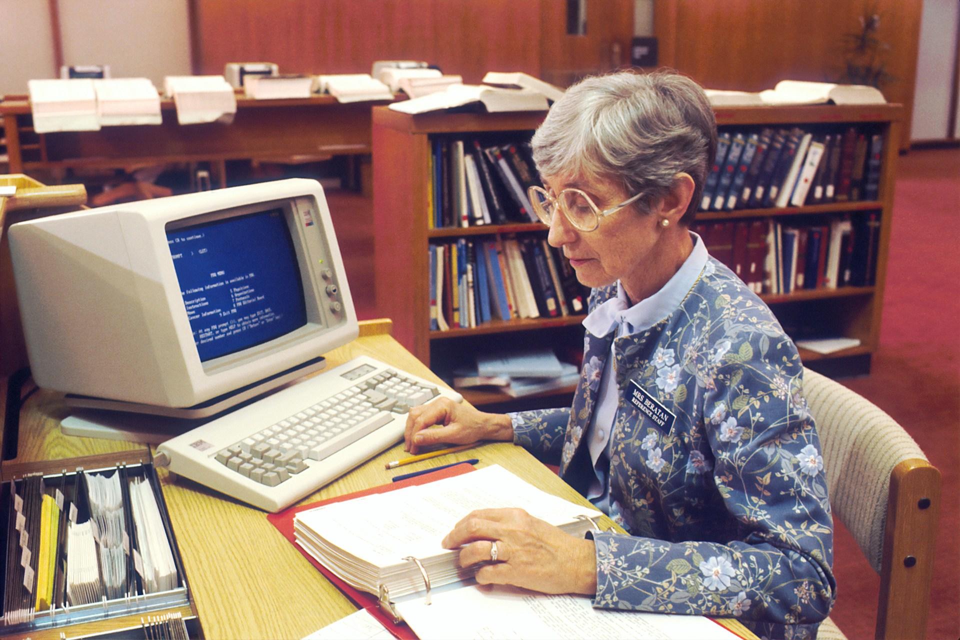 Eine ältere Person sitzt an einem altmodischen Computer und schaut in einen Ordner auf dem Tisch