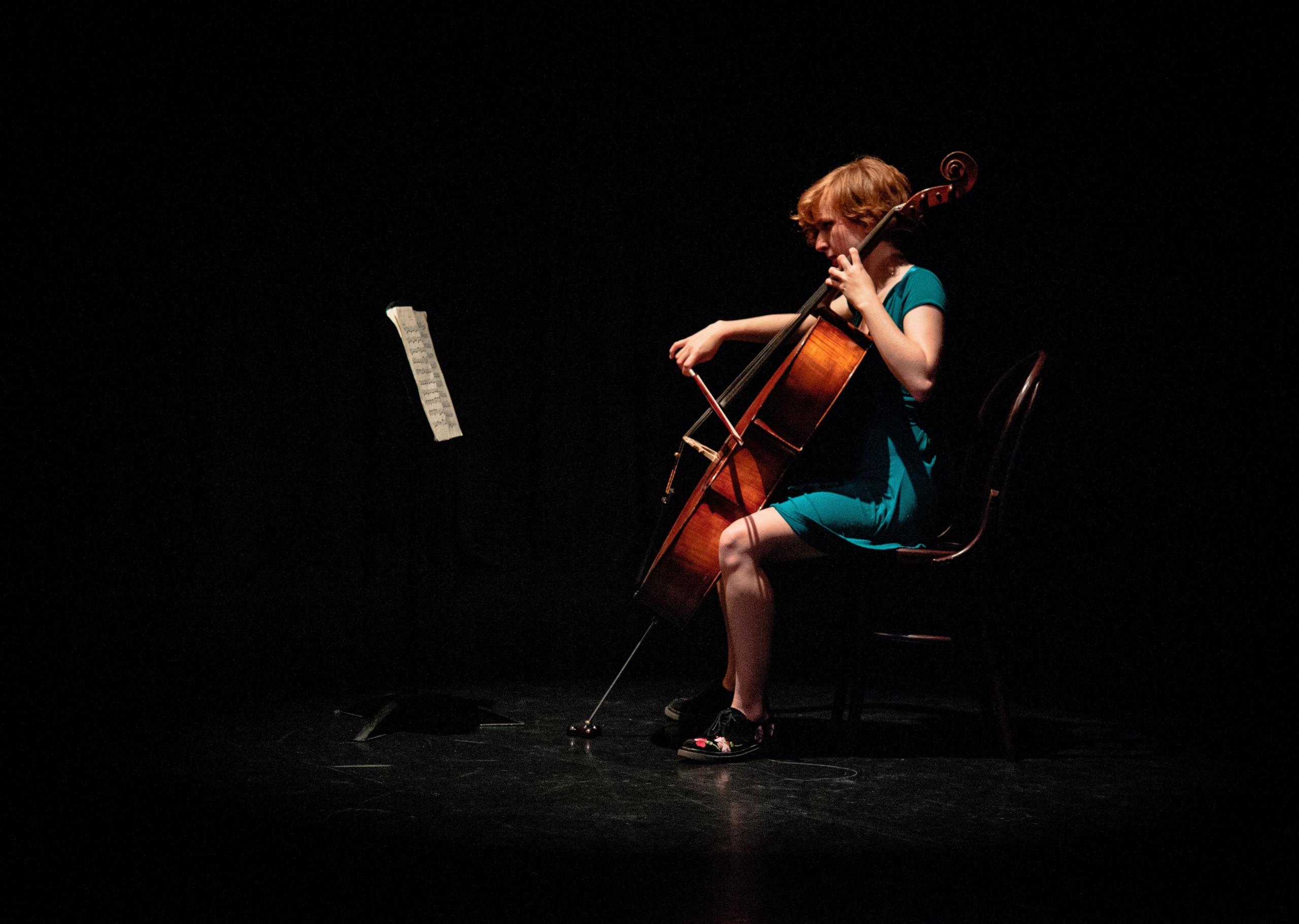 Cellistin mit Cello und Noten auf Stuhl vor schwarzem Hintergrund. So klappt es mit dem Cello lernen online.