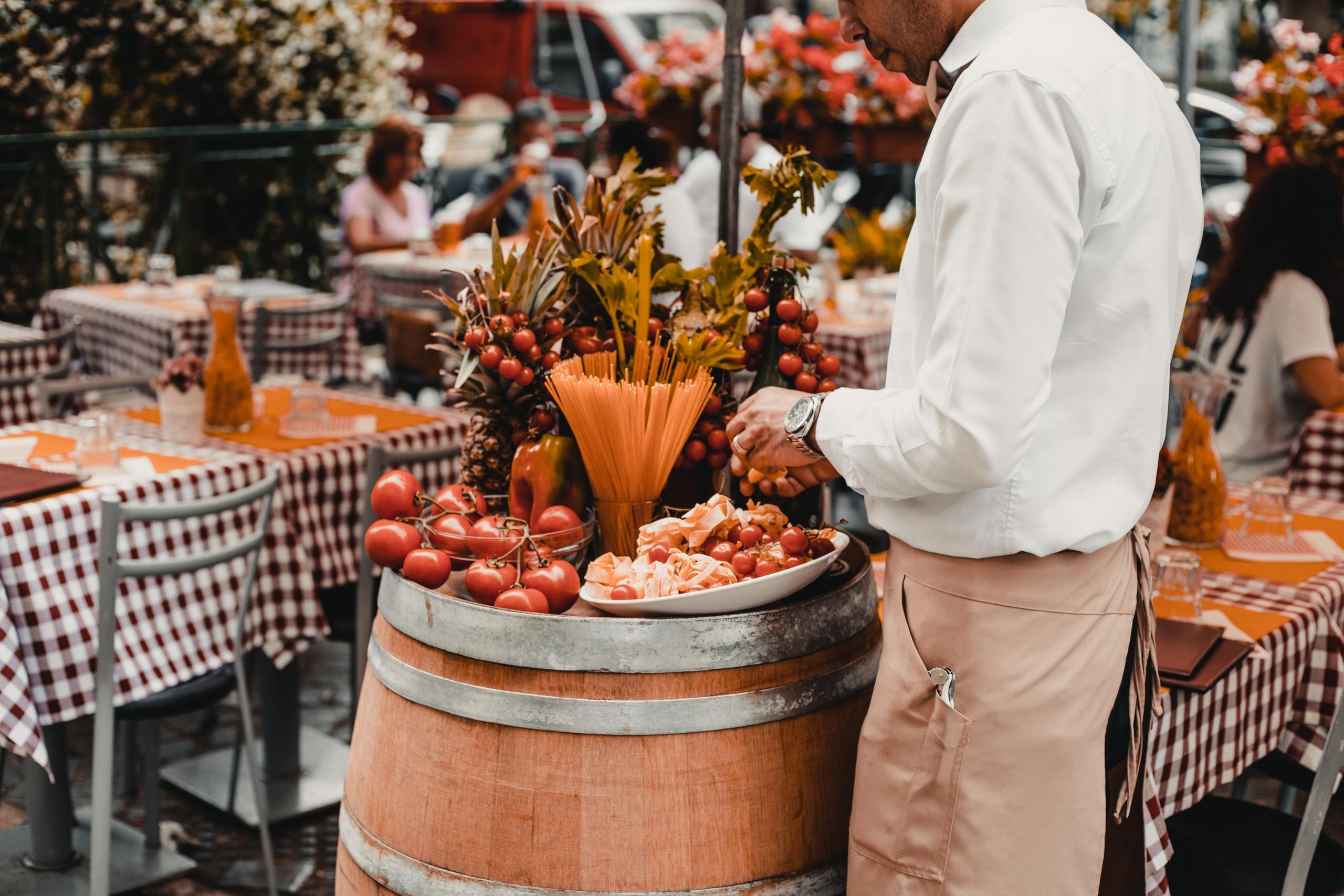 Kellner bereitet eine italienische Vorspeiseplatte mit Rohschinken und Tomaten auf einer Piazza mit Restaurantstühlen vor.