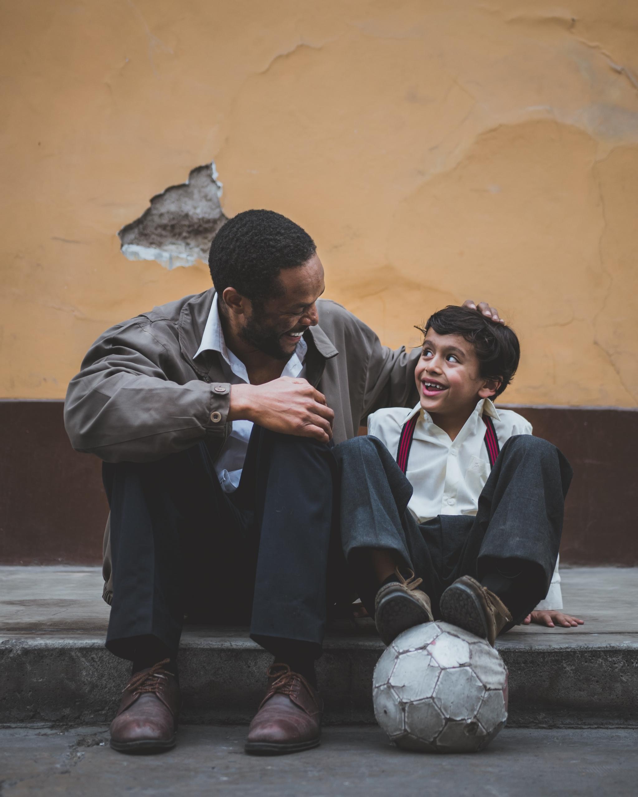 Ein Lehrer und ein kleiner Junge sitzen auf einem Bordstein, der Junge stützt seine Füße auf einen alten Fußball, beide sehen sich lachend an. Tutoring oder Nachhilfe hat viele Gesichter!