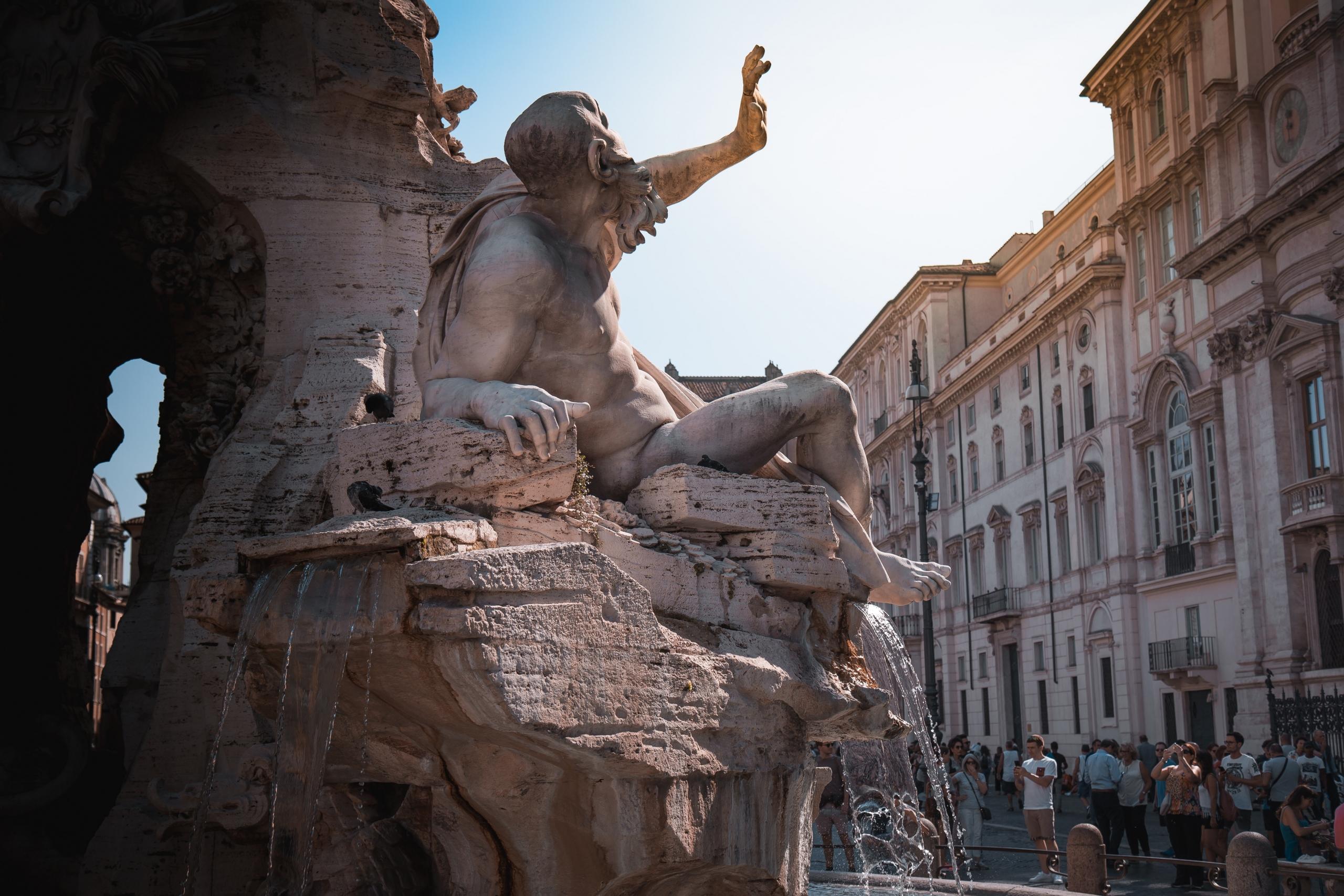 Ein antiker Brunnen mit einer männlichen Figur, die ihre Hand abwehrend von sich streckt, wahrscheinlich eine Szene aus römischer Mythologie? In römischer Kulisse lässt sich die Sonne genießen.