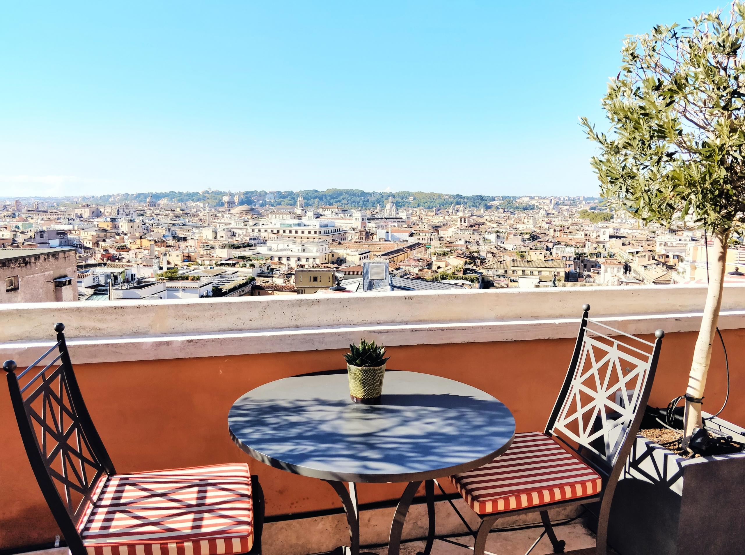 Blick über eine italienische Stadt unter blauem Himmel von einer terrakottafarbenen Terrasse aus, zwei ornamentierte Stühle und ein kleiner blauer Tisch darauf. Urlaub in Rom ist traumhaft und die verschiedenen Stadtteile eine Erkundung wert!