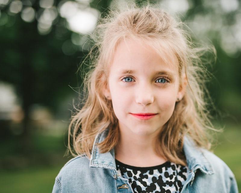 Ein blondes Mädchen mit hellen Augen in gepunktetem Oberteil und Jeanshemd schaut im Porträtformat in die Kamera. Fremdsprachen sind wichtig, denn sie öffnen den Blick der Kinder.