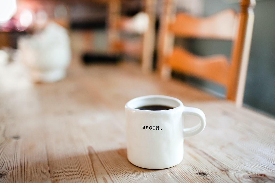 Auf einem rustikalen Holztisch steht eine weiße Tasse mit schwarzem Kaffee, auf der Tasse steht "Begin." - ein Neuanfang im Beruf klappt besser mit einer Beratung.