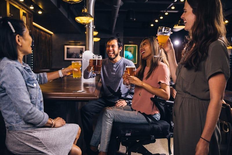 Eine gemischte Gruppe aus zwei Frauen und zwei Männern trinken Bier in einer urigen Bar und lachen miteinander. Auch abends treffen sich Kollegen noch auf einen Drink.