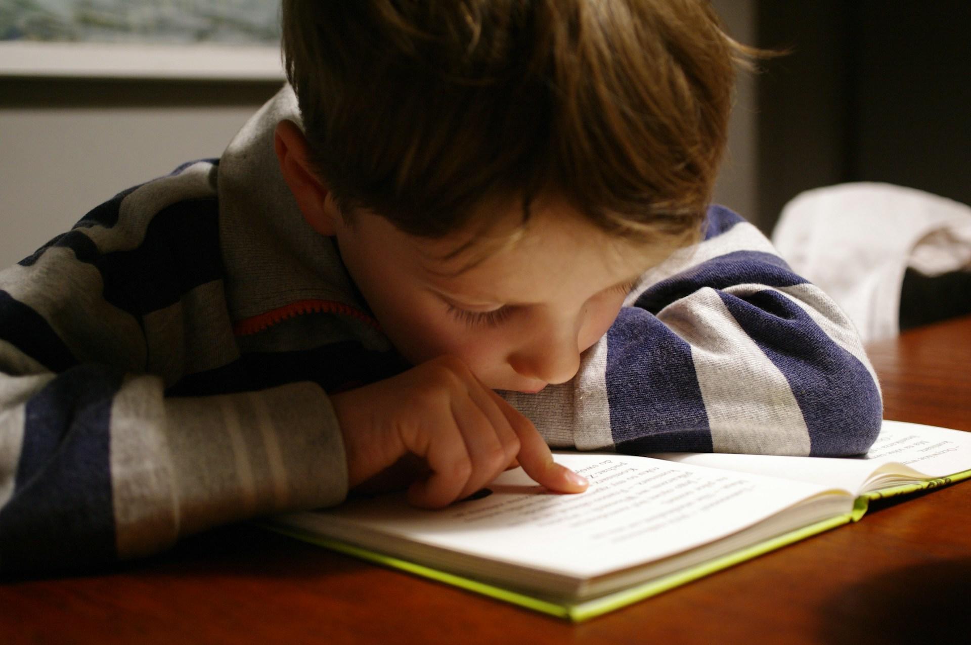 Ein kleiner Schüler liegt mit einem Arm auf einem Buch und liest mit einem Finger der anderen Hand einen Text.