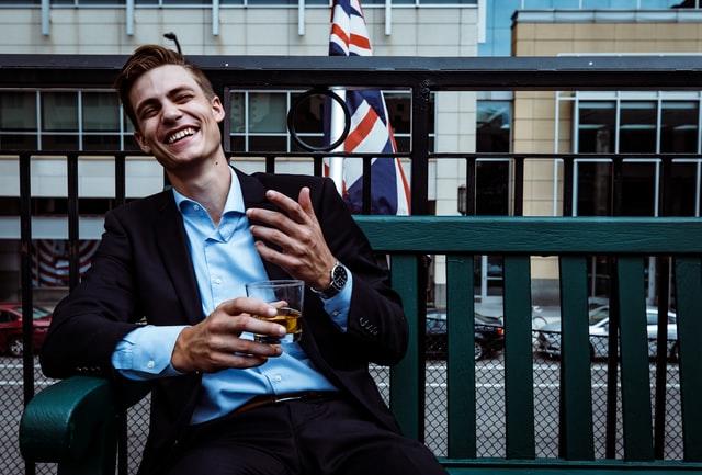 Ein junger Angestellter sitzt auf der Terrasse eines Pubs mit einem Drink in der rechten Hand vor einer einer britischen Fahne im Hintergrund. Willst du in England leben und arbeiten?