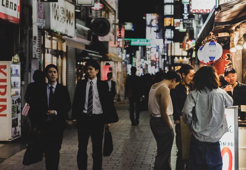 Am späten Abend laufen zwei japanische Büroangestellte nach der Arbeit durch eine belebte Straße mit Lampions und Lichtern, an der Seite stehen Menschen an einem Stand.