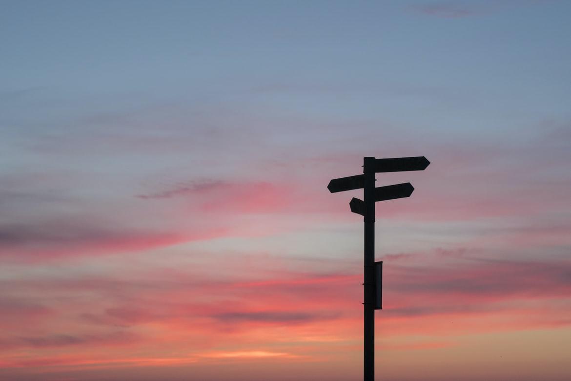 Vor einem in Pink und Orange getauchten Himmel zeichnen sich die dunklen Umrisse eines Schild mit Verweisen in verschiedene Richtungen ab. Berufswege und Karriereoptionen gibt es viele.