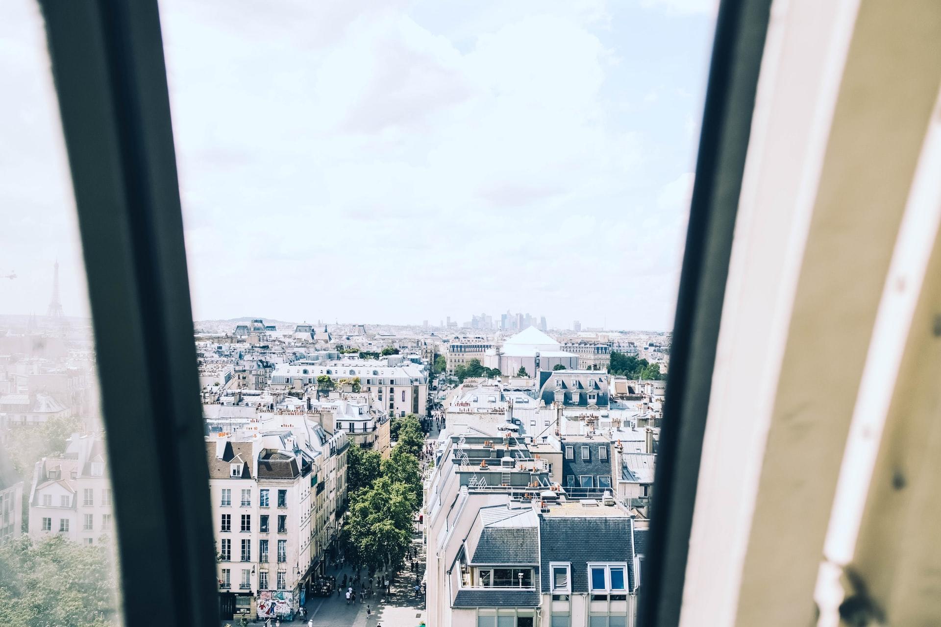 Blick durch ein geöffnetes Dachfenster auf Paris mit Eiffelturm im Dunst im Hintergrund. Bei einem französischen Unternehmen arbeiten und den Traum verwirklichen!