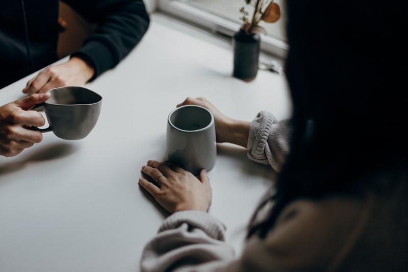 Zwei Menschen sitzen sich an einem Tisch gegenüber, jeder hält eine Tasse in der Hand. Über Probleme sprechen hilft!