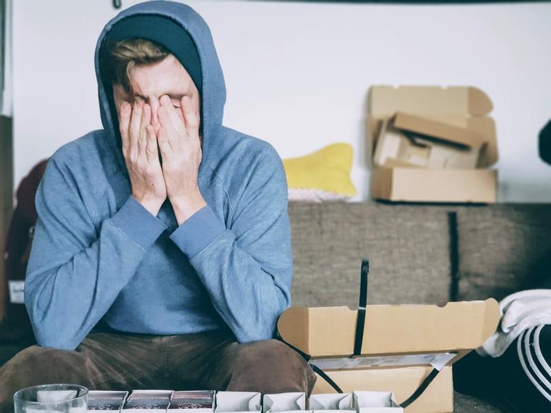 Ein junger Mann sitzt mit der Kapuze seines Hoodies hochgezogen inmitten von Kartons auf seinem Sofa und hält sich erschöpft die Hände vors Gesicht. Was tun gegen Burnout?