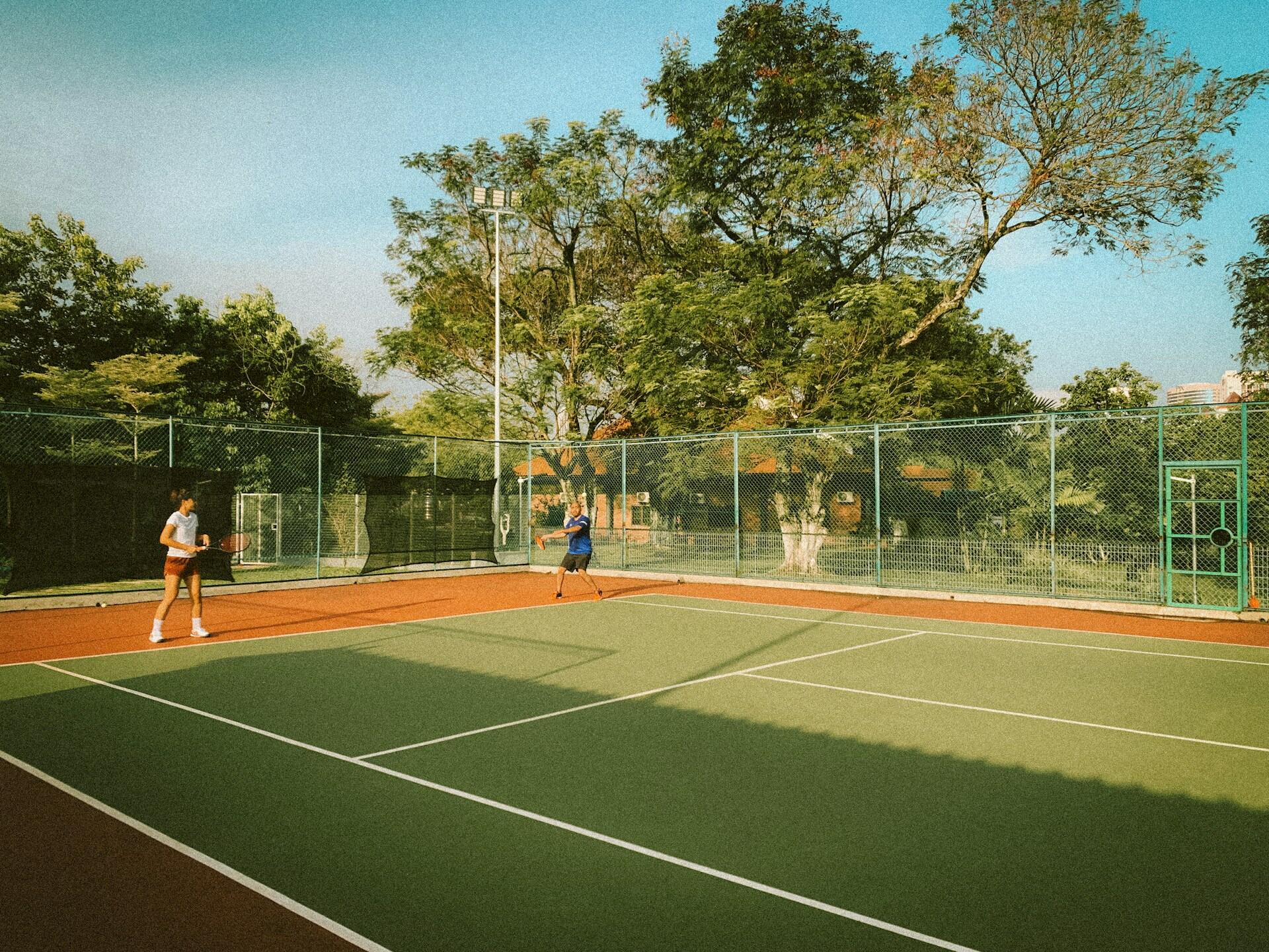 Zwei Personen spielen Tennis im Doppel auf dem Tennisplatz und einer macht den Aufschlag.