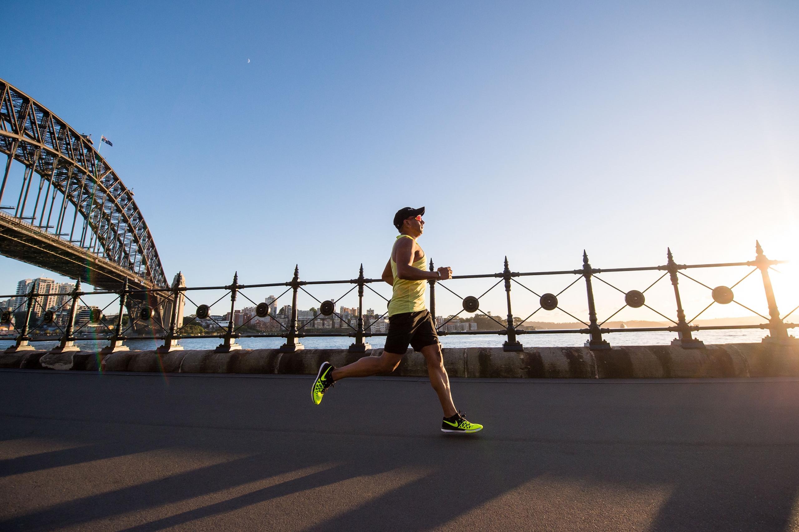 Ein Jogger auf dem asphaltierten Ufer eines Flusses, im Hintergrund eine Stahlbrücke und blauer Himmel. Ausdauer sollte zusätzlich zum Tennistraining trainiert werden. 
