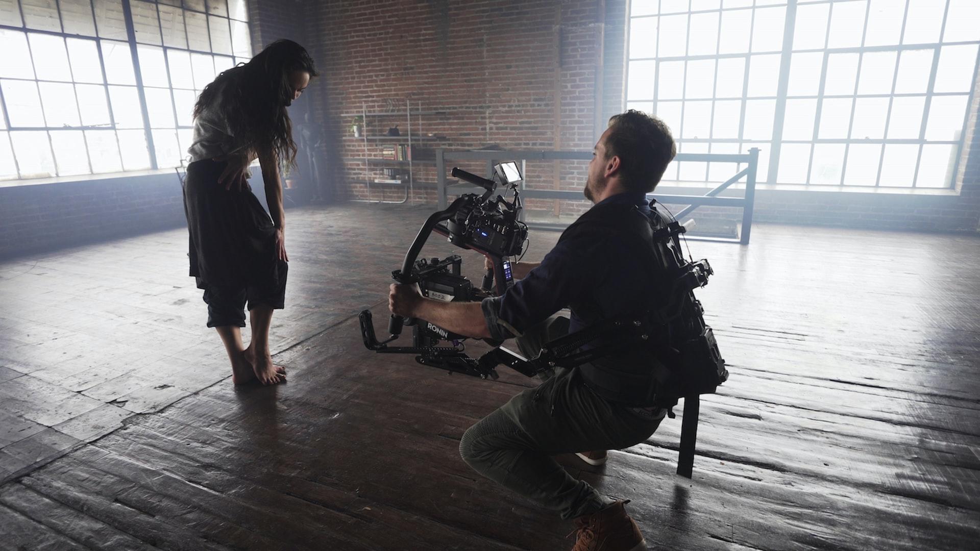 Eine junge Frau steht in einem leeren Saal und schaut runter gebeugt in die Kamera, welche vom Kameramann in einer unbequemen Haltung getragen wird. Method Acting fordert von den Darstellern alles!