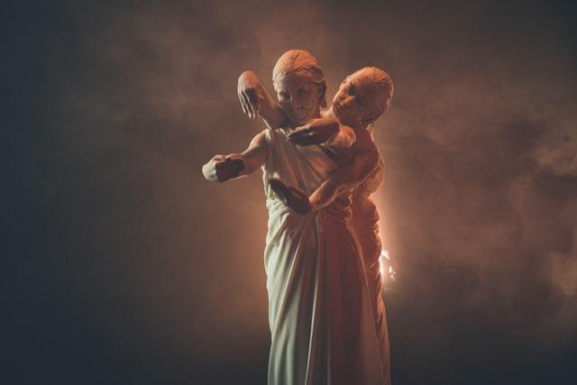 zwei Frauen stehen hinter einander auf der Bühne, in eine weisse Tunika gewickelt, weissen Lehm in Gesicht und Haar, mit den Armen nach vorne abstrakte Figuren machend. Theater kann viele Formen haben!