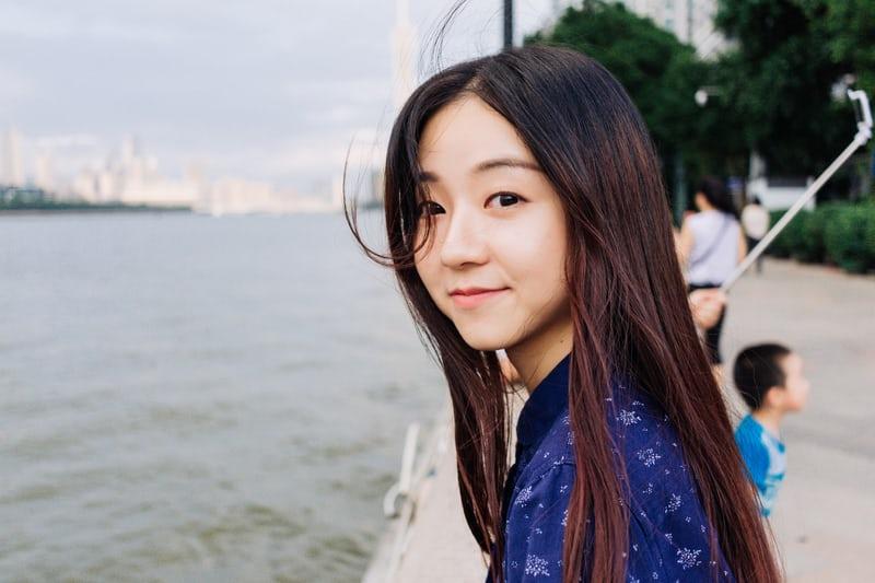 Ein junges koreanisches Mädchen blickt lächelnd seitlich in die Kamera, im Hintergrund ein urbaner Fluss.
