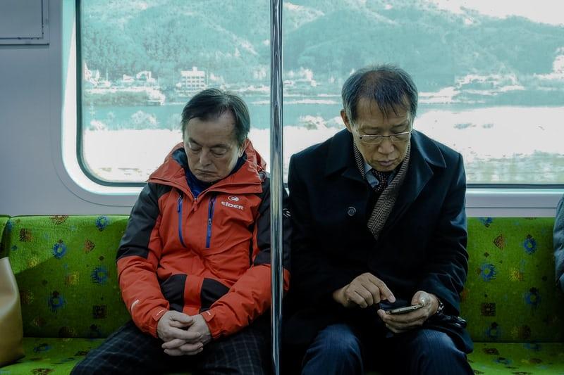 Zwei koreanische Männer sitzen in der Bahn, einer scheint zu schlafen, der andere guckt auf sein Handy. Im Hintergrund sieht man durchs Fenster die Landschaft. Koreaner achten auf einen höflichen Umgang.