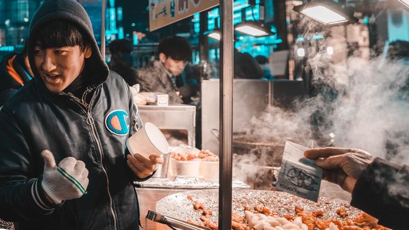 Ein Essensstand auf einem koreanischen Markt, im Vordergrund ein junger Mann.