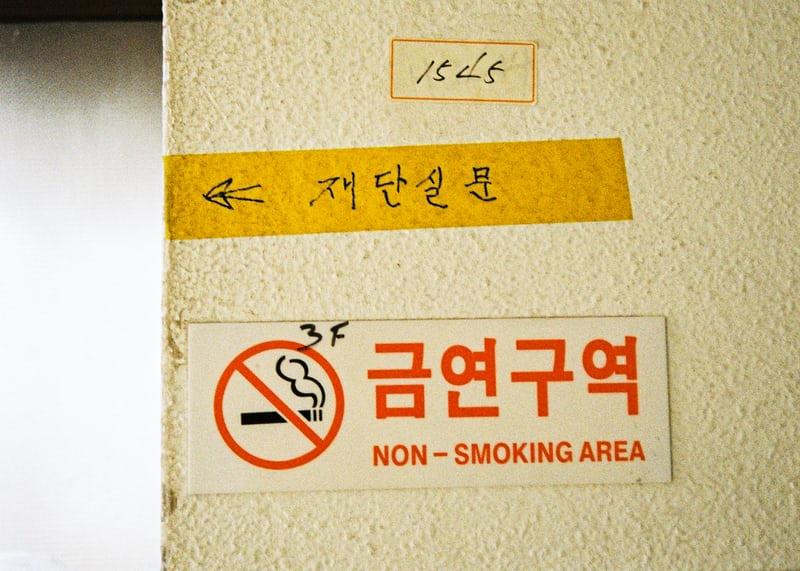 An einer Wand hängt ein Nichtraucherschild mit durchgestrichener Zigarette und koreanischen Schriftzeichen, daneben zwei weitere Aufschriften auf Koreanisch.