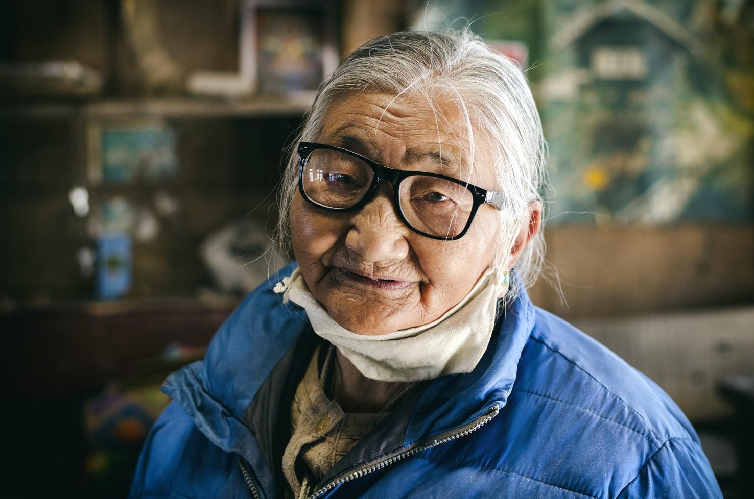 Eine alte asiatische Frau mit schwarzer Brille, Mundschutz unter dem Kinn und grauem Pferdeschwanz blickt in die Kamera.