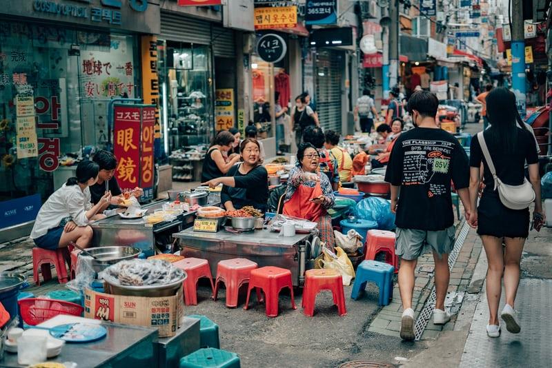Eine Marktszene in Südkorea. Frauen bieten in einer Strasse ihre Waren auf kleinen Tischen an, Menschen flanieren über den Markt.