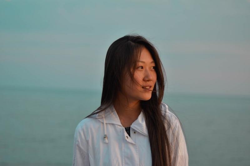 Eine junge, koreanische Frau mit langen Haaren blickt zur Seite, im Hintergrund das Meer oder ein See. In Stuttgart gibt es viele koreanische Muttersprachler.