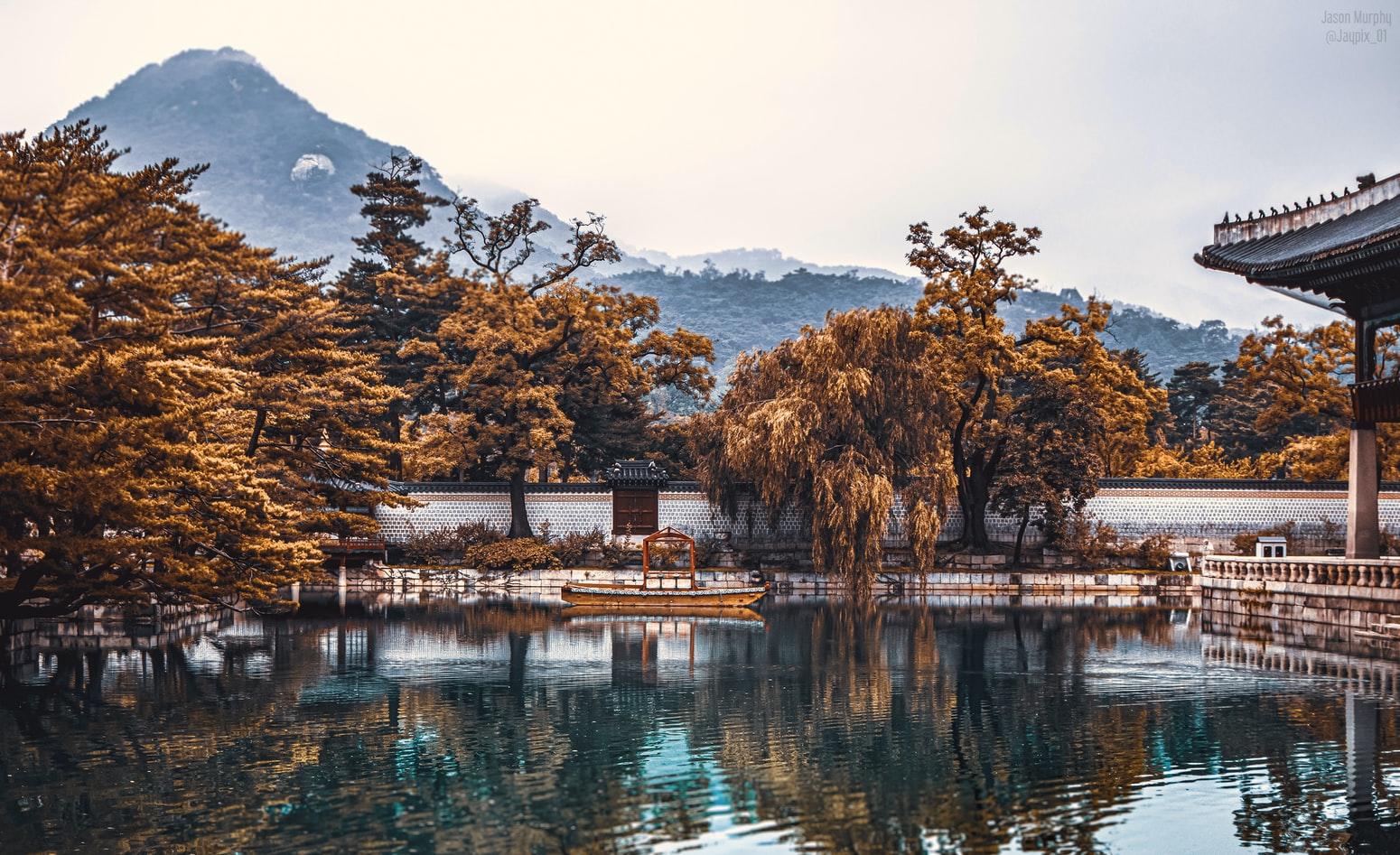 Im Vordergrund ein klarer See, dahinter am Ufer ein Tempel, herbstlich gelbe Bäume und im Hintergrund Berge. Südkorea ist ein exotisches Reiseland.