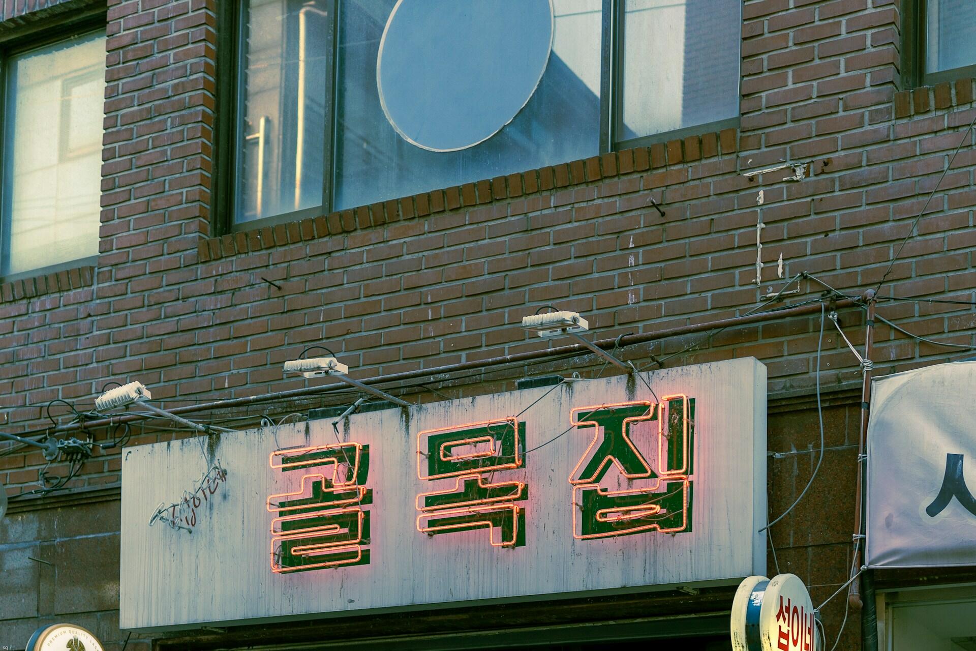 Schild mit koreanischer Schrift vor Backsteinmauer.