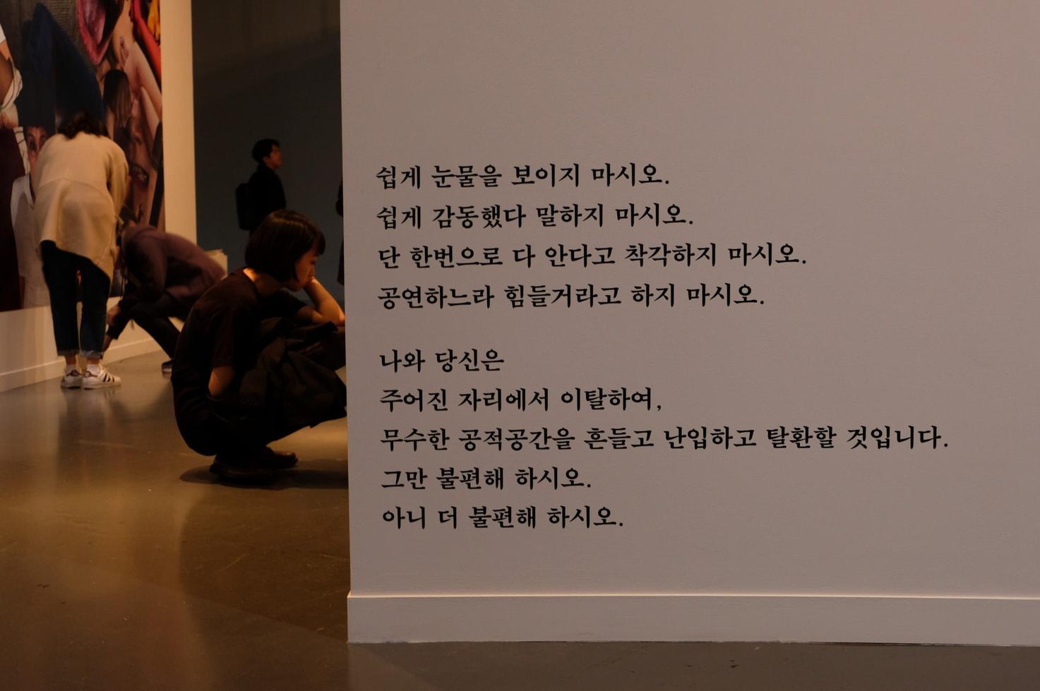 Ein Museum in Seoul. In Vordergrund eine weisse Wand mit schwarzen koreanischen Schriftzeichen, im Hintergrund Besucher vor einem Gemälde.