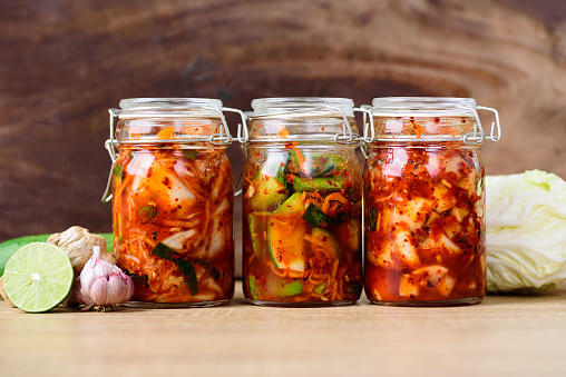 Drei grosse Einmachgläser mit verschiedenen Kimchi-Sorten. Daneben ein Kohlkopf, Knoblauch und eine aufgeschnittene Limette.