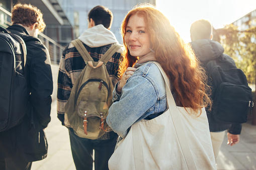 Eine junge, rothaarige Studentin in Jeansjacke und mit grosser Tasche über der Schulter dreht sich um und lächelt. Im Hintergrund laufen drei junge Männer mit Rucksack weiter auf ein Gebäude zu. Auf dem Weg zum Institut für Koreanistik.