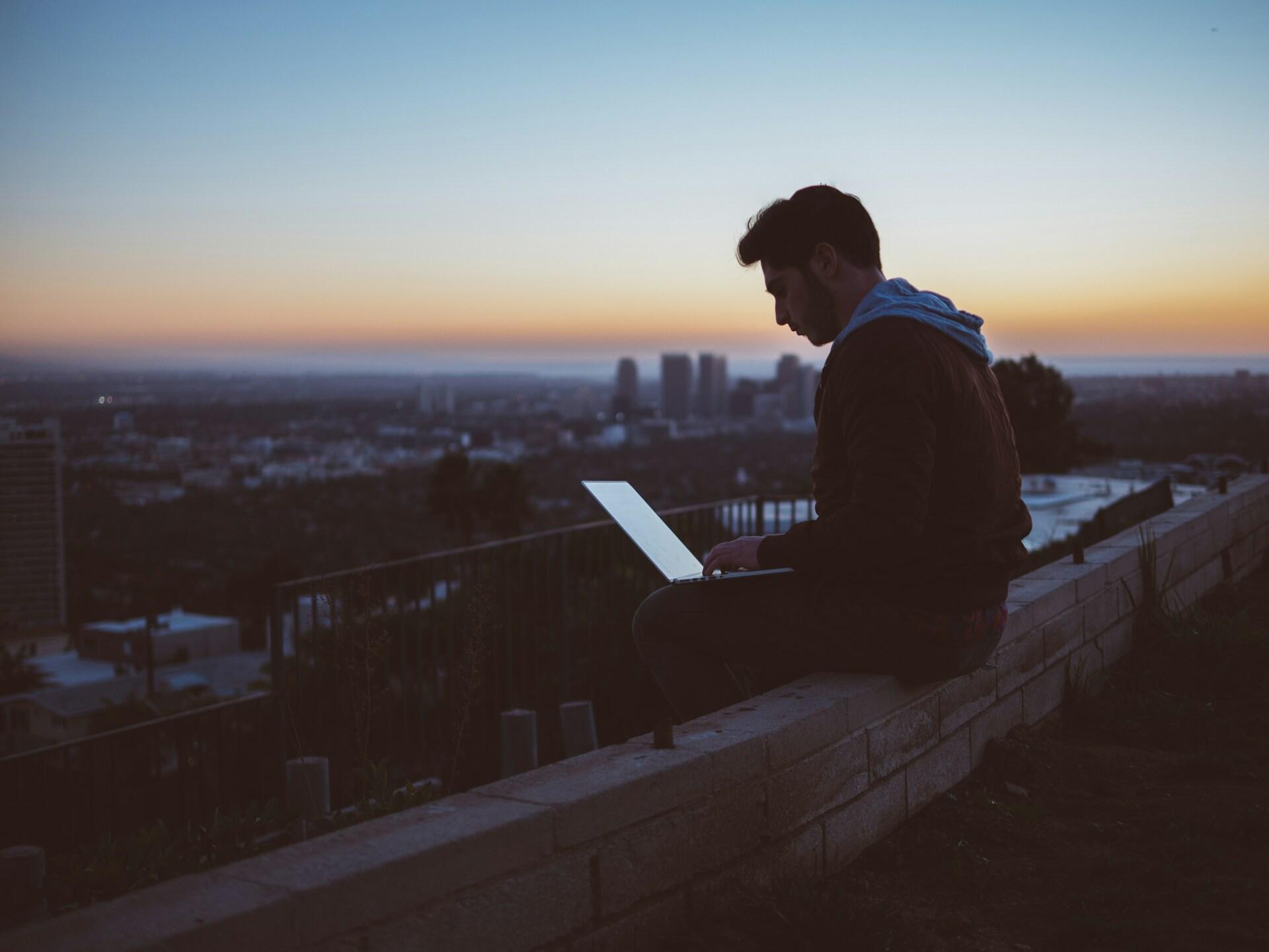 Ein Mann sitzt mit Laptop auf den Knien im Sonnenuntergang draußen auf einer Mauer. Im Hintergrund ist eine Großstadt zu erkennen.