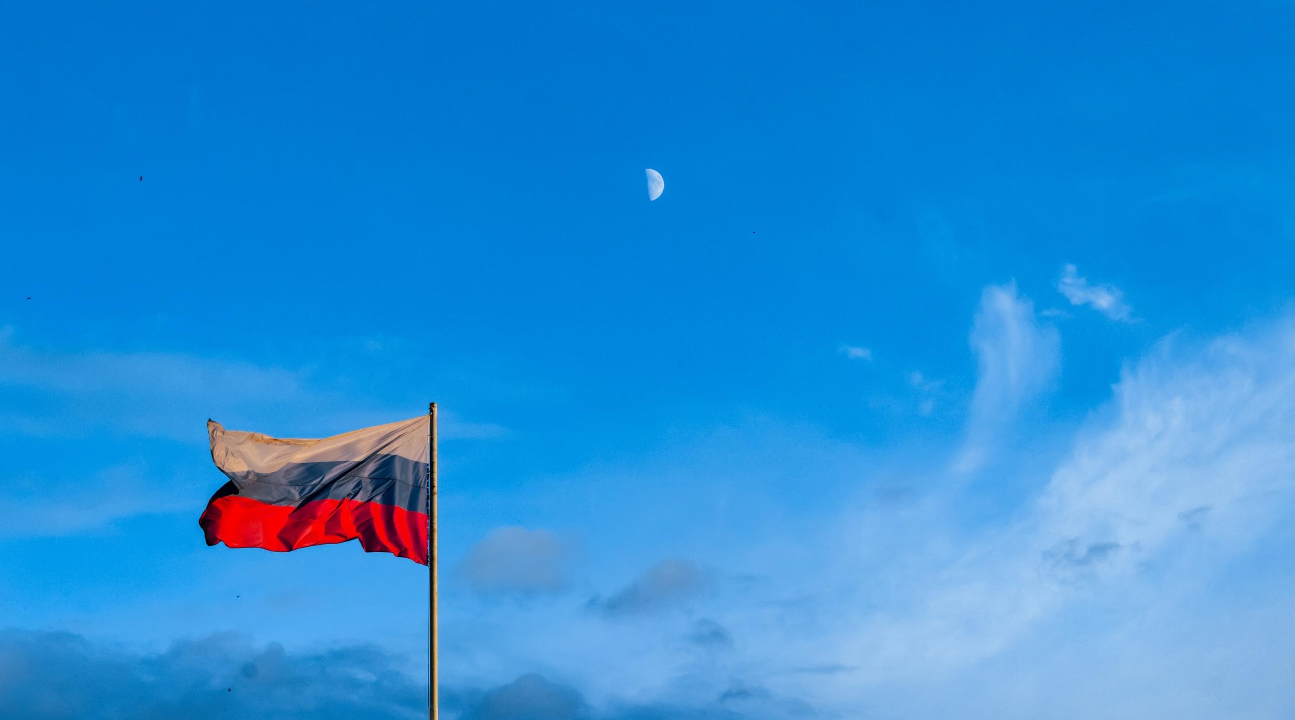 Die Flagge der russischen Föderation weht vor blauem Himmel, der Mond steht am Himmel, obwohl es Tag ist. Wie wäre es mit einer Reise nach Russland?