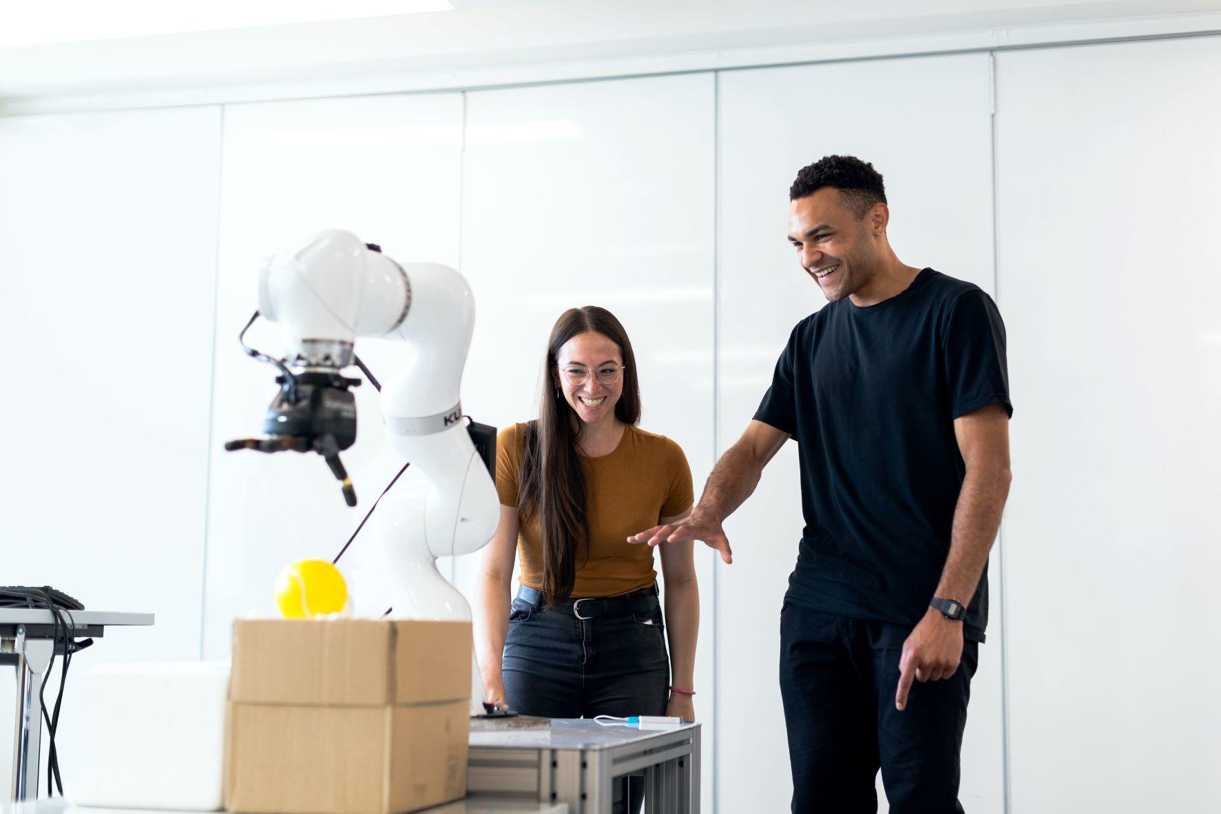 Eine junge Frau und ein junger Mann stehen vor einem Tisch mit einem Roboter, während der Mann auf den Roboter zeigt und beide lachen. Erst wenn man weiß, wie und wofür der Roboter programmiert wurde, weiß man, was er alles kann. 